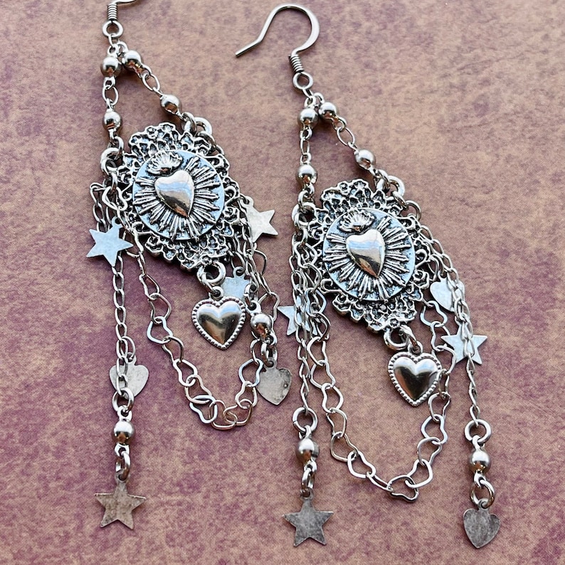 Puede incluir: Pendientes de plata con un dije de coraz&oacute;n, estrellas y cadenas. El dije de coraz&oacute;n tiene un dise&ntilde;o detallado y una corona en la parte superior.