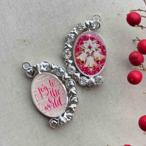 May include: Two silver-toned Christmas ornaments. One features the phrase "Joy to the World" in pink script over sheet music. The other has a red background with gold bells, white poinsettias, and snowflakes. Both have heart and crystal accents.
