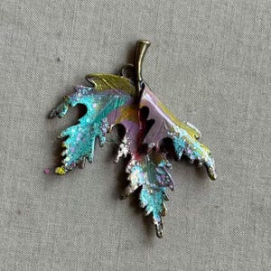 May include: A colorful leaf pendant with a bronze stem and edges. The leaf is painted with shades of teal, purple, yellow, and green, with glitter accents. The pendant is likely made of metal.