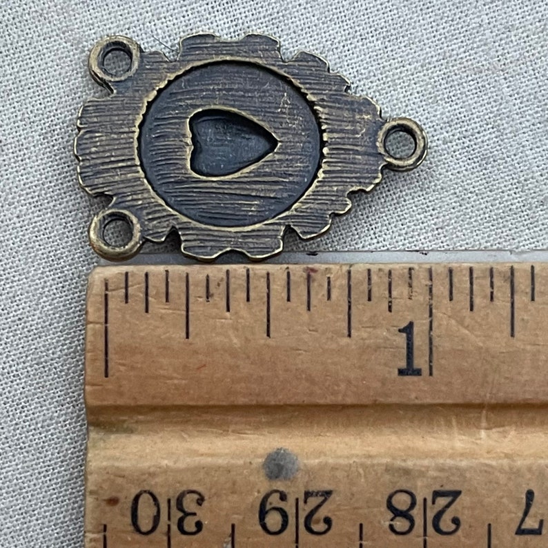 Puede incluir: Un dije de metal de color bronce con un recorte en forma de coraz&oacute;n en el centro. El dije tiene dos bucles a cada lado, lo que lo hace adecuado para la elaboraci&oacute;n de joyas u otros proyectos de artesan&iacute;a. El dije mide aproximadamente 2,5 cm de largo.