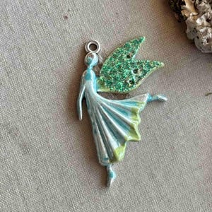 May include: A silver charm featuring a fairy with green glitter wings and a blue and green dress. The fairy is in a dancing pose with one leg raised.
