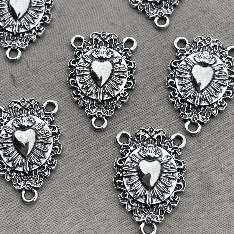 May include: Five silver-toned metal charms with a heart design. Each charm features a heart with a crown on top, surrounded by a decorative floral pattern. The charms have three jump rings for attaching to jewelry.
