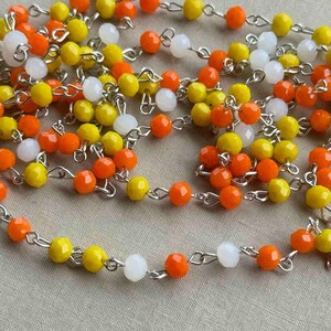 May include: Close-up of beaded jewelry strands. The strands feature faceted beads in orange, yellow, and white, strung on silver-colored chains. The beads are evenly spaced, creating a textured and colorful pattern.