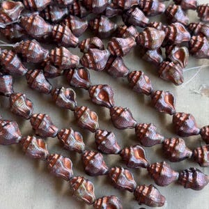 May include: A collection of metallic copper-toned beads shaped like small seashells, strung together on thin white thread. Each bead features a textured, ridged surface with a glossy finish, displaying variations of bronze and brown hues throughout.