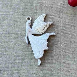 May include: White metal fairy charm with wings outstretched and a flowing skirt. The charm has a distressed finish and a loop for hanging.