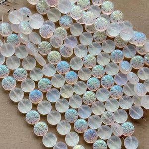 May include: A collection of round, translucent glass beads featuring an iridescent finish and a pressed snowflake pattern on the surface. The beads are arranged in a loose cluster, displaying a pale, milky white color with shifting rainbow reflections.