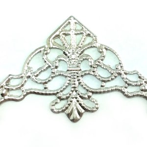 May include: Silver filigree corner embellishment with a diamond-shaped design in the center. The embellishment has a delicate, intricate pattern and is perfect for adding a touch of elegance to crafts and projects.