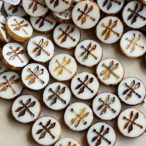 May include: A collection of round, white beads with dragonfly designs. Each bead features a dragonfly in shades of brown and gold, with a brown border. The beads are likely for jewelry making or crafts.