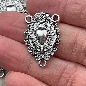May include: Silver-toned metal charm featuring a heart with a crown and rays of light. The charm has two jump rings for attaching to jewelry or other crafts.