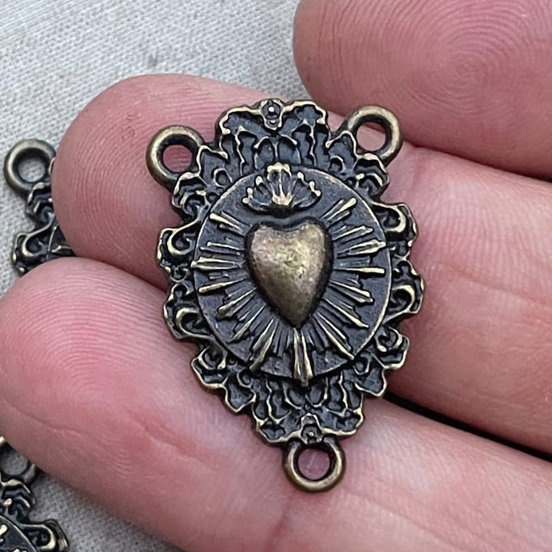 Puede incluir: Un dije de metal dorado con forma de coraz&oacute;n coronado, rodeado de un c&iacute;rculo de rayos. El dije tiene un borde decorativo y dos anillos de salto para sujetarlo a un collar o pulsera.