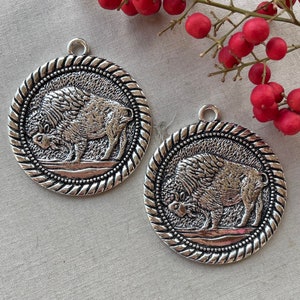 May include: Two silver-colored round pendants with a detailed bison design. Each pendant has a rope-like border and a small loop at the top for hanging. The pendants are on a neutral background.