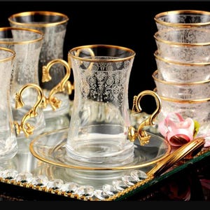 May include: A set of six clear glass teacups with gold rims and intricate floral designs. The cups are arranged on a mirrored tray with a gold trim and crystal accents.