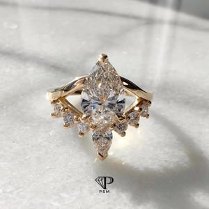 May include: A gold ring with a large, pear-shaped diamond as the centerpiece. The ring features smaller diamonds around the main stone and along the band, creating a sparkling, elegant design. The ring is set on a white surface.