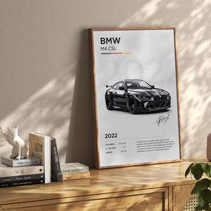 May include: A black and white poster featuring a black BMW M4 CSL with the text "BMW M4 CSL 2022" and technical specifications. The poster has a wood frame.