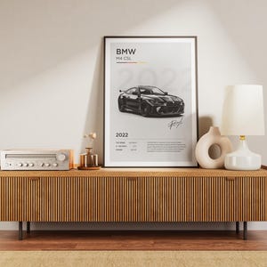 May include: A black and white poster with a black BMW M4 CSL car on a white background. The poster has the text "BMW M4 CSL 2022" and a signature at the bottom.