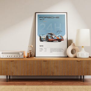 K&ouml;nnte beinhalten: Ein gerahmtes Poster mit blauem Hintergrund, auf dem ein Vintage-Porsche 917-Rennwagen abgebildet ist. Das Poster tr&auml;gt den Text "PORSCHE 917 24H 1970". Das Poster ist auf einem Holzregal mit einer Stereoanlage und einer Lampe ausgestellt.