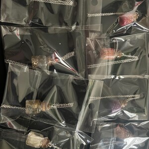 Op de afbeelding: Acht transparante glazen flesjes met zilveren kettingen en verschillende gekleurde glitters erin. De flesjes zijn individueel verpakt in doorzichtige plastic zakjes.