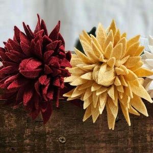 Puede incluir: Primer plano de dos flores de fieltro. Una es de color burdeos intenso y la otra amarillo claro. Las flores tienen múltiples pétalos puntiagudos, que se asemejan a las dalias. Las flores hechas a mano están sobre una superficie de madera.