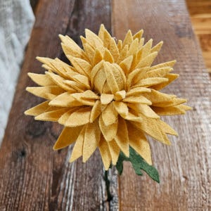 May include: A handmade, yellow felt chrysanthemum flower with a green stem and leaves. The flower has multiple layers of petals, creating a full, textured appearance. The felt flower is a decorative item.