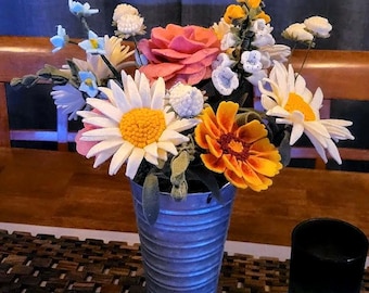 May include: A colorful bouquet of felt flowers in a galvanized metal vase. The arrangement includes daisies, roses, and other blooms in shades of pink, yellow, orange, and white. The vase is cylindrical and has a ribbed texture.