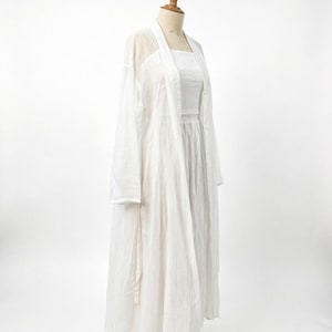 May include: A white, full-length dress and a matching long, open-front cardigan are displayed on a mannequin. The dress has a square neckline and a fitted bodice. The cardigan is sheer and lightweight, creating a layered look. The outfit is made of a soft, flowing fabric.