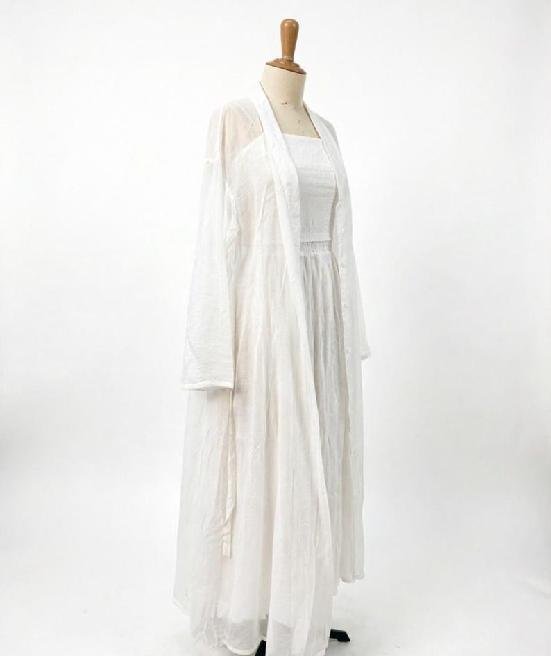 May include: A white, full-length dress and a matching long, open-front cardigan are displayed on a mannequin. The dress has a square neckline and a fitted bodice. The cardigan is sheer and lightweight, creating a layered look. The outfit is made of a soft, flowing fabric.