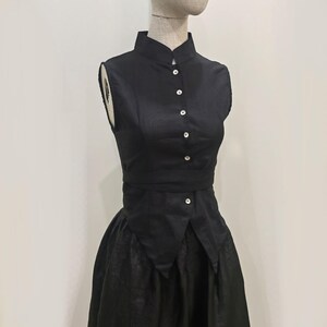 Könnte beinhalten: Ein schwarzes ärmelloses Kleid mit Stehkragen und eng anliegendem Oberteil. Das Kleid hat kleine, weiße Knöpfe und einen ausgestellten Rock. Das Kleid wird auf einer Schaufensterpuppe präsentiert.