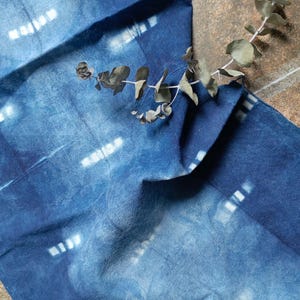 Könnte beinhalten: Ein blauer Stoff mit einem weißen Batik-Muster. Der Stoff ist gefaltet und ein Zweig getrockneter Eukalyptusblätter liegt darauf.