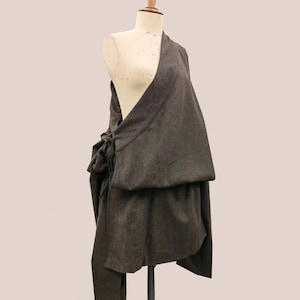 May include: A dark gray wrap-style garment draped on a mannequin. The fabric appears to be a soft wool or wool blend. The wrap is tied at the side with a bow, and the design features a layered effect. The garment is a neutral color.