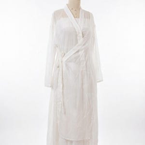 May include: A white wrap dress with long sleeves, displayed on a mannequin. The dress features a V-neck and a tie closure. The fabric is lightweight and semi-transparent, with a layered skirt. The background is plain white.