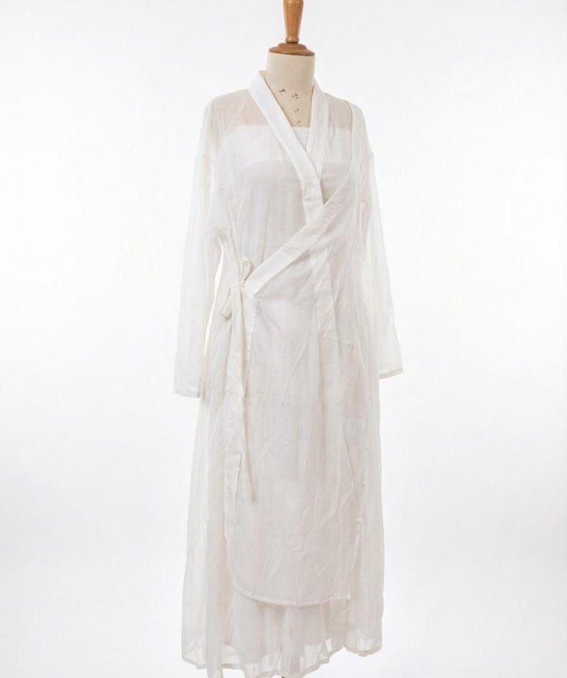 May include: A white wrap dress with long sleeves, displayed on a mannequin. The dress features a V-neck and a tie closure. The fabric is lightweight and semi-transparent, with a layered skirt. The background is plain white.