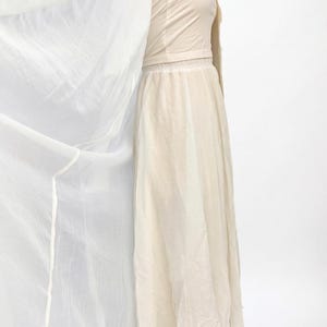 May include: A cream-coloured dress and matching jacket. The dress features a long, flowing skirt and a fitted bodice. A sheer, white fabric is draped to the side, adding movement and texture. The outfit is made of lightweight fabric, suitable for warmer weather.