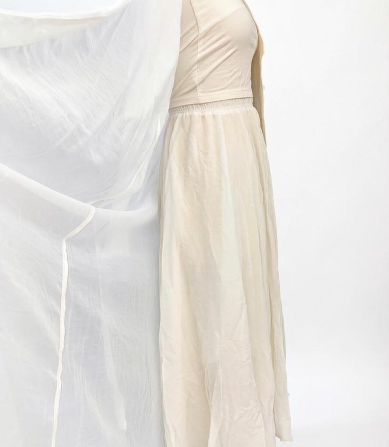 May include: A cream-coloured dress and matching jacket. The dress features a long, flowing skirt and a fitted bodice. A sheer, white fabric is draped to the side, adding movement and texture. The outfit is made of lightweight fabric, suitable for warmer weather.