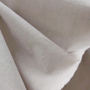 Puede incluir: Primer plano de una tela lisa de color blanco roto con una superficie ligeramente texturizada. La tela está doblada y muestra el tejido del material.