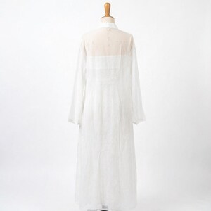 May include: A long, white, sheer dress with long sleeves is displayed on a mannequin. The dress has a high collar and a semi-transparent yoke across the shoulders and upper back. The fabric appears lightweight and flowing.