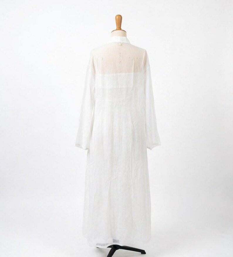 May include: A long, white, sheer dress with long sleeves is displayed on a mannequin. The dress has a high collar and a semi-transparent yoke across the shoulders and upper back. The fabric appears lightweight and flowing.