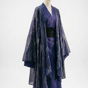 Puede incluir: Una bata estilo kimono azul oscuro con un patrón plateado, superpuesta a un atuendo azul a juego. La bata tiene mangas anchas y se asegura con un cinturón negro. El atuendo se exhibe en un maniquí sobre un fondo blanco.