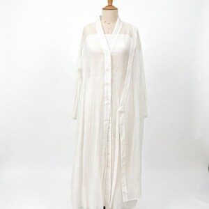 May include: A white, floor-length dress and matching long-sleeved overcoat displayed on a mannequin. The dress has a square neckline and the overcoat has a button-up front and a tie belt. The fabric appears lightweight and semi-sheer.