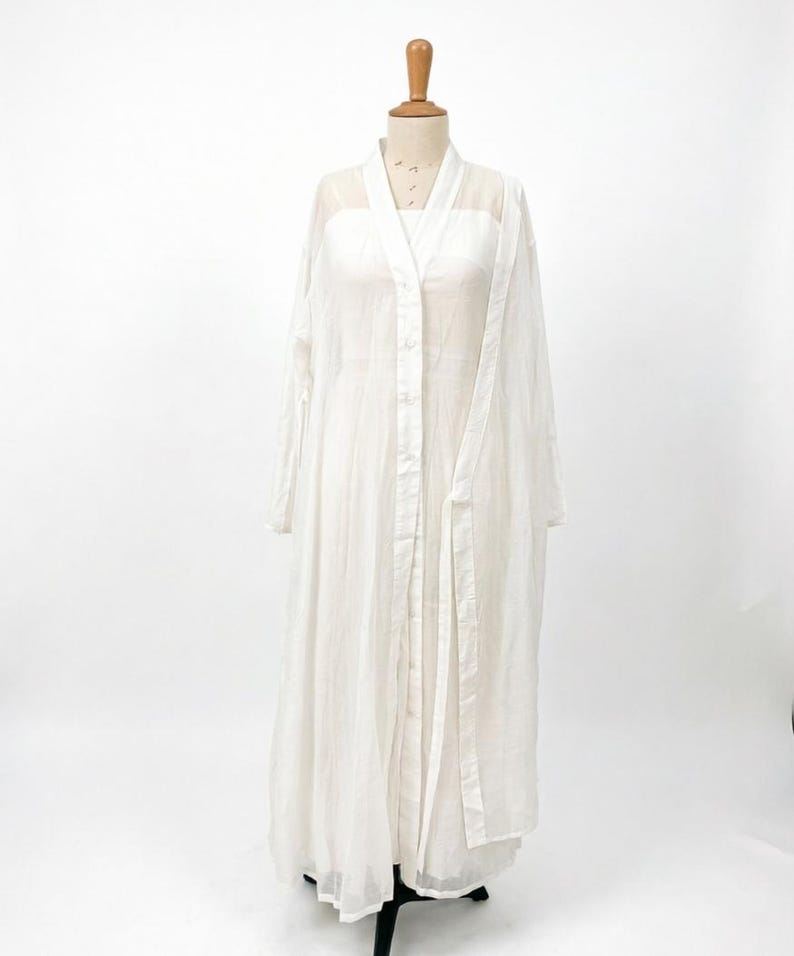 May include: A white, floor-length dress and matching long-sleeved overcoat displayed on a mannequin. The dress has a square neckline and the overcoat has a button-up front and a tie belt. The fabric appears lightweight and semi-sheer.