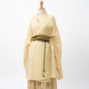 May include: A light yellow linen kimono-style top and skirt ensemble on a mannequin. The top has a wrap-style closure and long sleeves, cinched with a green sash. The skirt is a similar color with a floral pattern.
