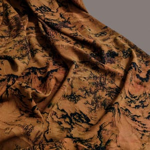 May include: Close-up of a fabric with a landscape design. The fabric has a warm, earthy tone with a detailed pattern of mountains, trees, and other natural elements in black. The fabric is draped, showing its texture and flow.