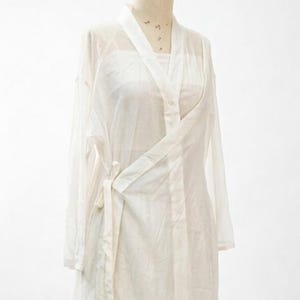 May include: A white, sheer wrap top with long sleeves and a tie fastening. The top is made of a lightweight fabric and has a draped design. The garment is displayed on a mannequin.