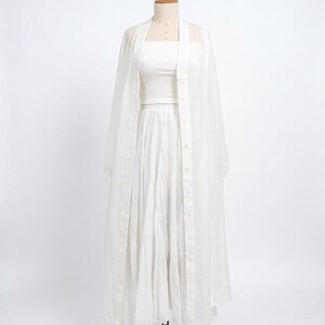 May include: A white three-piece outfit on a mannequin. The ensemble includes a tube top, a pleated skirt, and a sheer, button-down duster. The duster has long sleeves and a collarless design. The outfit is displayed against a plain white background.