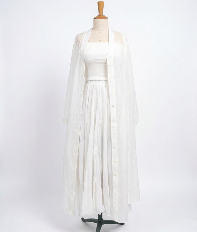 May include: A white three-piece outfit on a mannequin. The ensemble includes a tube top, a pleated skirt, and a sheer, button-down duster. The duster has long sleeves and a collarless design. The outfit is displayed against a plain white background.