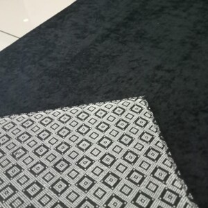 May include: A black, textured rug with a corner of a patterned fabric. The fabric has a repeating geometric design in black and white. The rug appears to be made of a soft material.
