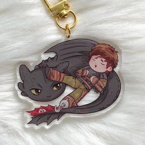 May include: A clear acrylic keychain featuring a cartoon illustration of a black dragon and a person with brown hair. The person is lying on the dragon. The keychain has a gold-colored clasp.