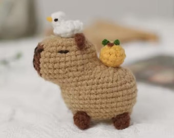 May include: A brown crocheted capybara plush toy with a white crocheted bird on its head and a yellow crocheted fruit on its back.