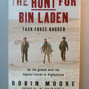 Puede incluir: Portada del libro "The Hunt for Bin Laden" de Robin Moore. El título está en letras rojas grandes. La portada presenta a tres soldados caminando por un paisaje desértico. También se ve el texto "Task Force Dagger".