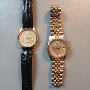 Puede incluir: Dos relojes de pulsera con cajas y esferas doradas. El reloj de la izquierda tiene una correa de cuero negra y el reloj de la derecha tiene una correa de metal de dos tonos, plata y oro. Ambos relojes muestran las palabras "De Anza Valley College" en la esfera.