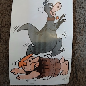 May include: A vintage iron-on sticker featuring Fred Flintstone and Dino from the cartoon series The Flintstones. The sticker shows Fred spinning in a barrel while Dino runs around him. The sticker is from 1976 and is made by Hanna-Barbera Productions, Inc.